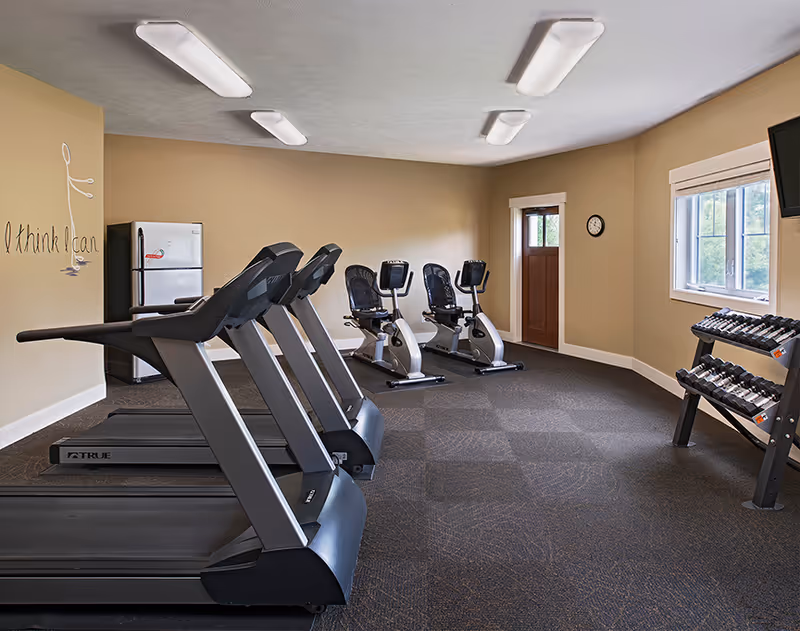 A fitness room with three treadmills on the left, two stationary exercise bikes in the back, and a rack of dumbbells on the right. The room has beige walls, a window, a wall clock, a door, and a refrigerator in the corner. On the left wall, there is a motivational phrase that reads 'I think I can' with a simple drawing of a person lifting weights.