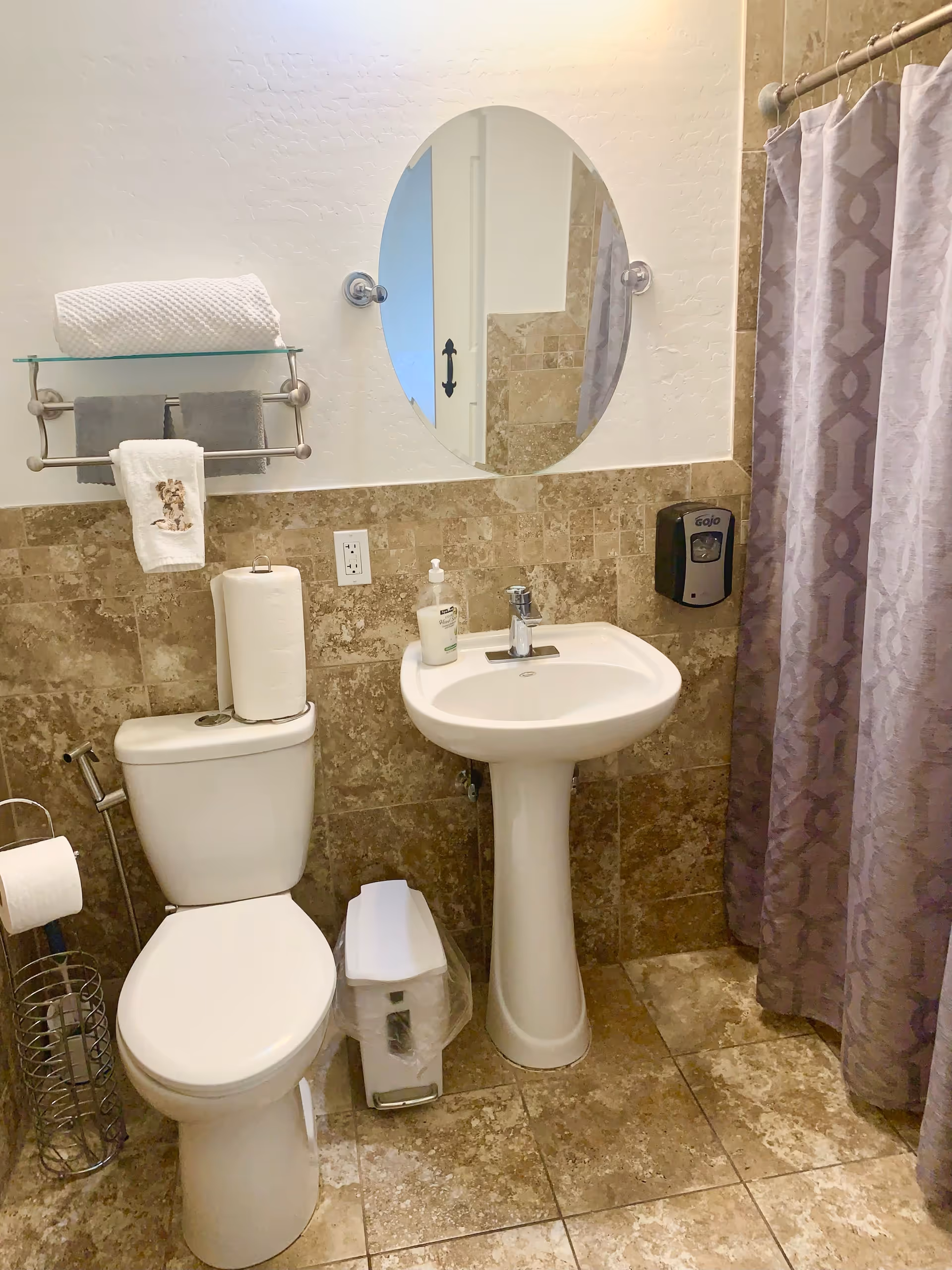 A clean bathroom with a white toilet, a pedestal sink with a soap dispenser, a small trash can, and a shower with a patterned curtain. There is a round mirror above the sink, a towel rack with folded towels, and beige tiled walls and floor.