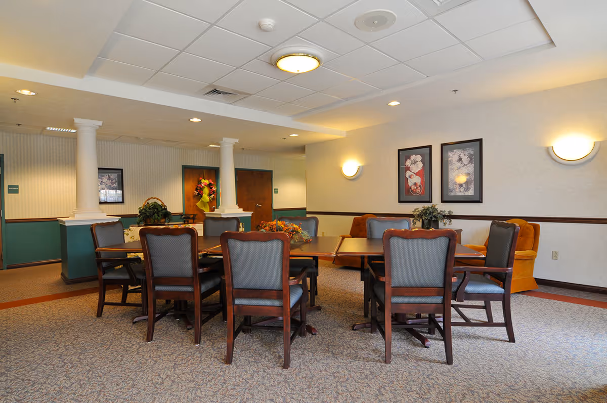 A well-lit common area with a large wooden table surrounded by eight upholstered chairs. The room features beige walls with two framed floral artworks, two orange armchairs, decorative plants on the table and in the background, and white columns near the back wall with wooden doors.