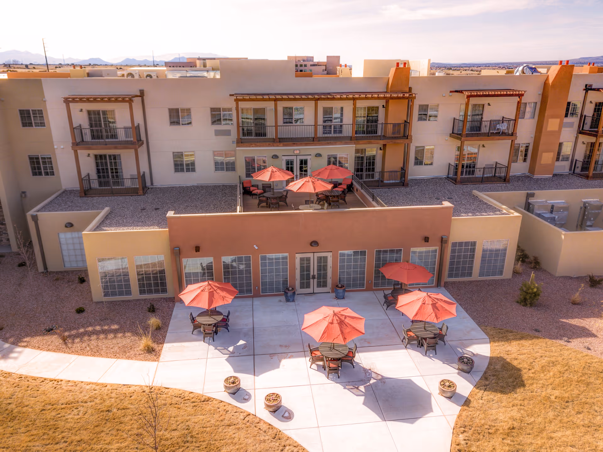 Outdoor patio area of a senior living facility with several round tables shaded by red umbrellas. The patio is adjacent to a beige building with large windows and balconies on the upper floors. The surrounding area includes a walkway and dry landscaping with grass and small plants.