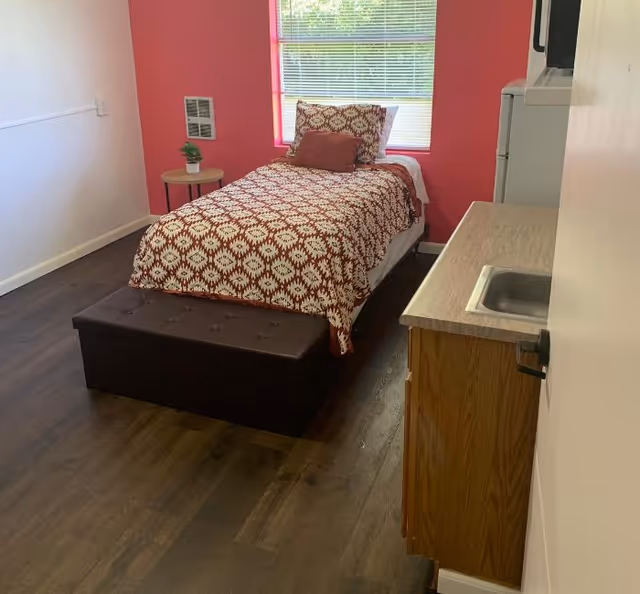 A small room with a single bed covered in a red and white patterned bedspread, a matching pillow, and a smaller red cushion. There is a window with blinds behind the bed, a small round side table with a potted plant next to the bed, and a dark brown storage bench at the foot of the bed. On the right side of the room, there is a kitchenette area with a countertop, a small sink, and a microwave on top of a mini refrigerator. The walls are white except for one red accent wall behind the bed, and the floor is dark wood.