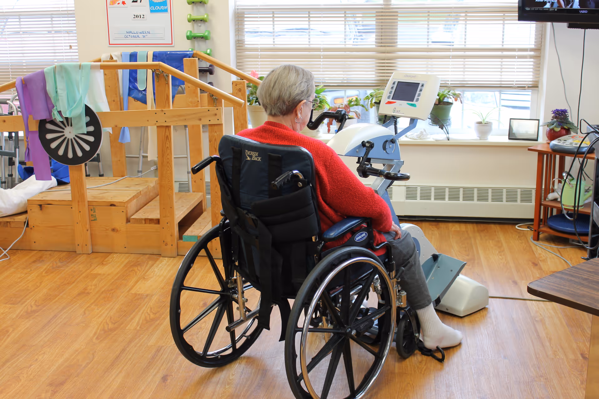 An elderly person in a wheelchair using a stationary exercise machine in a rehabilitation room with wooden flooring, handrails, exercise bands, and potted plants on the windowsill.