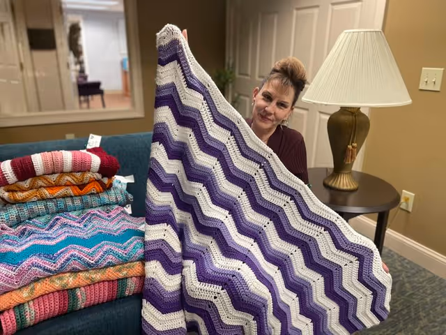 A woman sitting on a blue couch indoors, holding up a large crocheted blanket with purple and white zigzag stripes. Several other colorful crocheted blankets are stacked on the couch beside her. A table with a lamp is visible in the background.
