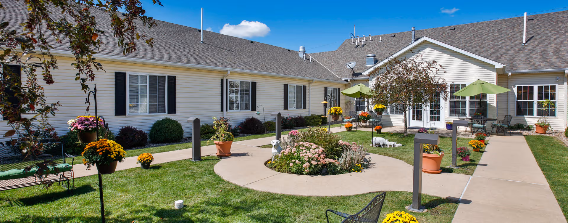 Outdoor courtyard area at Bickford of Peoria featuring a circular flower bed with various colorful flowers, surrounded by a paved walkway. There are several potted plants, green umbrellas shading seating areas, and a white building with multiple windows and black shutters in the background under a clear blue sky.