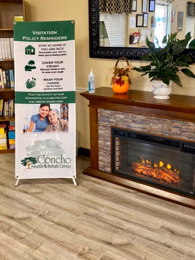 Interior view of a room in Concho Health and Rehabilitation Center showing a wooden electric fireplace with a stone facade. On top of the fireplace mantel, there is a small decorative pumpkin with fall-themed decorations and a potted green plant. To the left of the fireplace, there is a standing sign with visitation policy reminders including instructions to stay home if sick, cover coughs, and wash hands. A bottle of hand sanitizer is placed on the mantel. A large mirror hangs above the fireplace, reflecting part of the room.