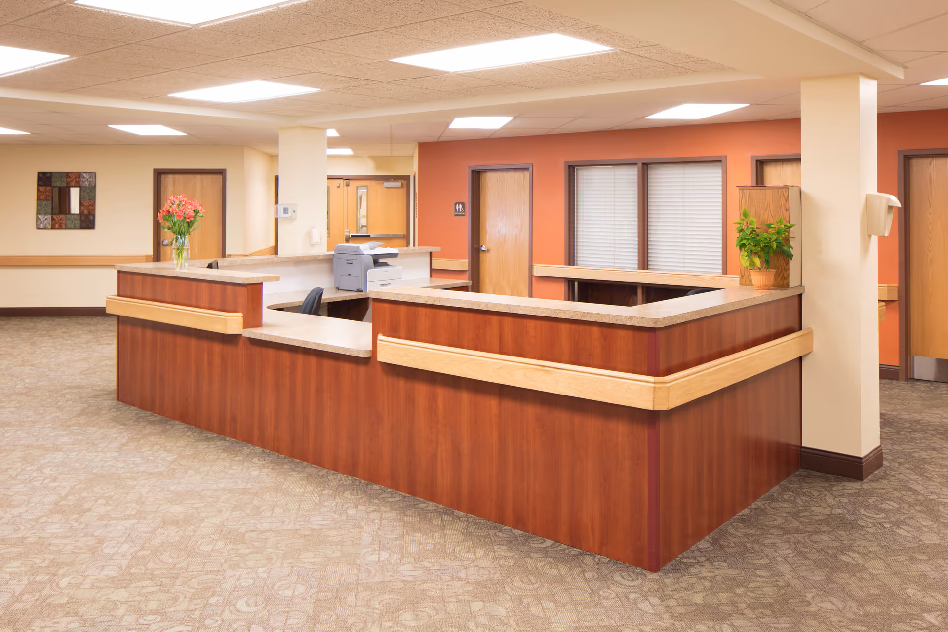 Reception area in a nursing and rehabilitation facility with a large wooden desk, a vase of flowers, a potted plant, and several closed wooden doors in the background.