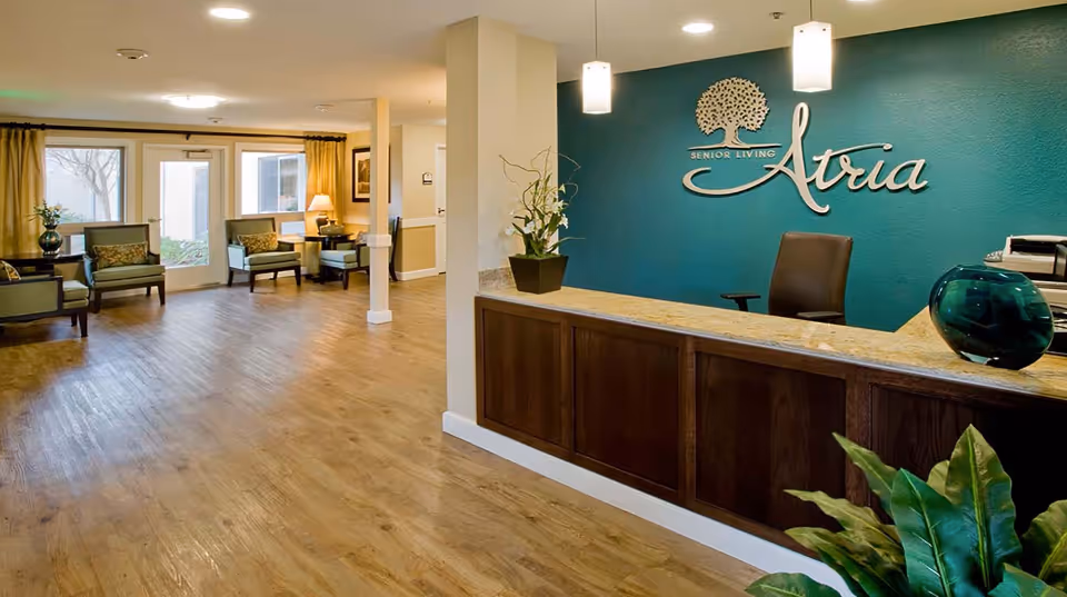 Reception area of Atria Sunnyvale senior living facility with a wooden front desk, a teal accent wall featuring the Atria logo, pendant lights above the desk, and a seating area with chairs and a table near large windows.