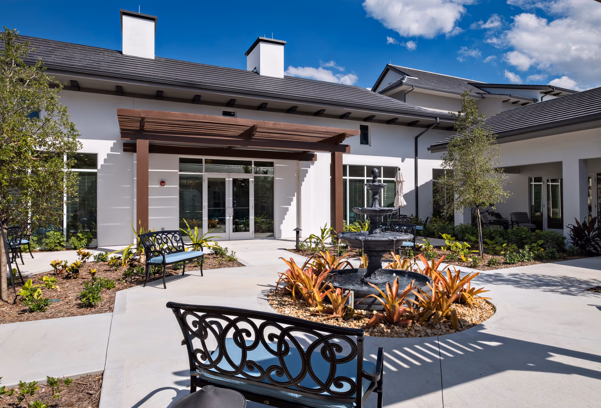 Outdoor courtyard area of a senior living facility with a black metal fountain surrounded by plants and two black metal benches with blue cushions. The building has white walls, large windows, and a wooden pergola over the entrance. The sky is blue with some clouds.