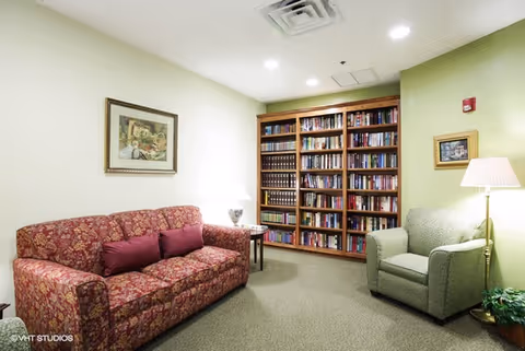 A cozy living room area with a red patterned sofa and two purple cushions, a green armchair, a wooden bookshelf filled with books, two table lamps, and framed artwork on the walls.