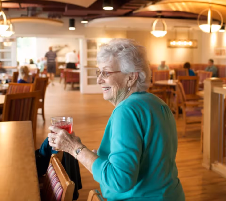 An elderly woman wearing a green sweater smiles while holding a glass in a communal dining room.