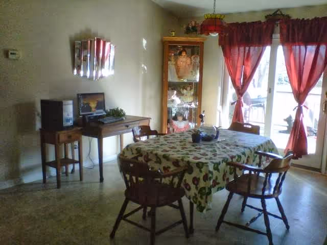 Dining room with a table covered by a patterned tablecloth surrounded by wooden chairs, a display cabinet, a small desk with a computer, and red curtains over glass doors.