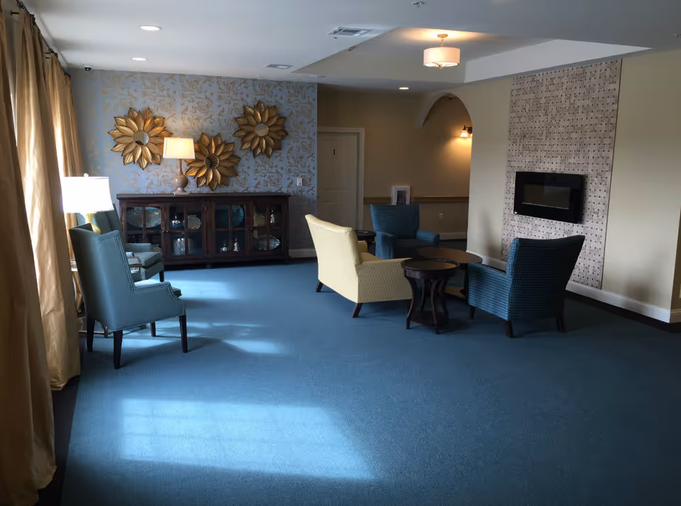 A cozy living room area with blue carpet and light beige walls. The room features a wall-mounted electric fireplace with a decorative tile surround. There are four upholstered chairs arranged around two small round tables. On the left side, there is a wooden cabinet with glass doors, topped with a lamp and three gold sunburst mirrors on the wall above. Large windows with beige curtains allow natural light to enter the room.