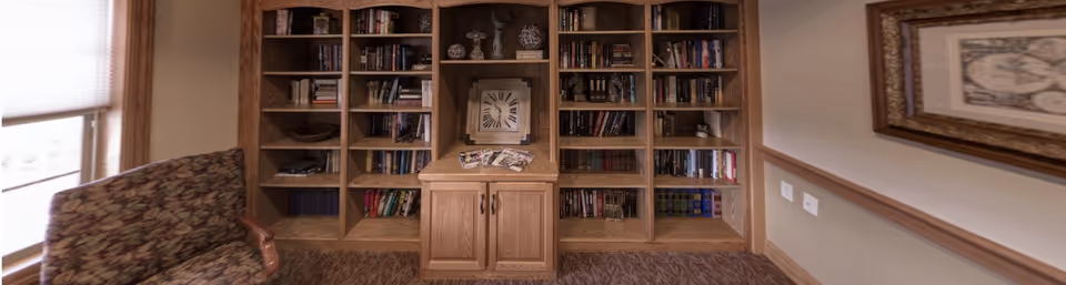 A cozy interior room featuring a large wooden bookshelf filled with books and decorative items, a patterned upholstered armchair on the left, and a framed picture hanging on the right wall.