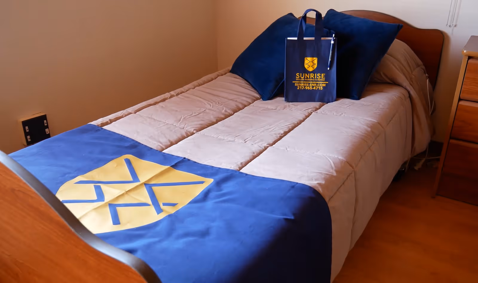 A neatly made single bed with beige bedding and a blue blanket featuring a yellow emblem at the foot. Two dark blue pillows are placed at the head of the bed, along with a blue tote bag displaying the Sunrise Skilled Nursing and Rehab logo, website, and phone number. The room has wooden flooring and a wooden nightstand beside the bed.