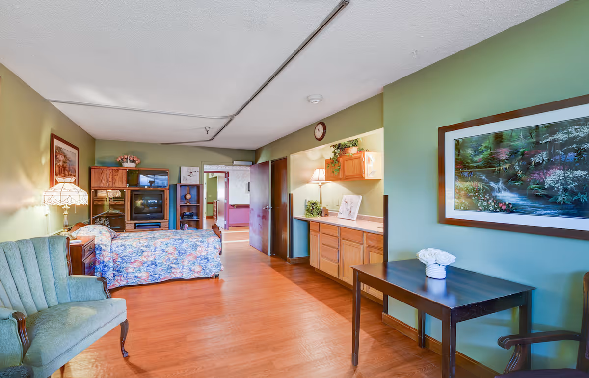 A cozy room with green walls and wooden flooring featuring a floral-patterned bed, a green upholstered armchair, a wooden entertainment center with a TV, a table with a white flower vase, and a framed nature painting on the wall. There is also a countertop with a lamp and decorative items, and an open door leading to another room.