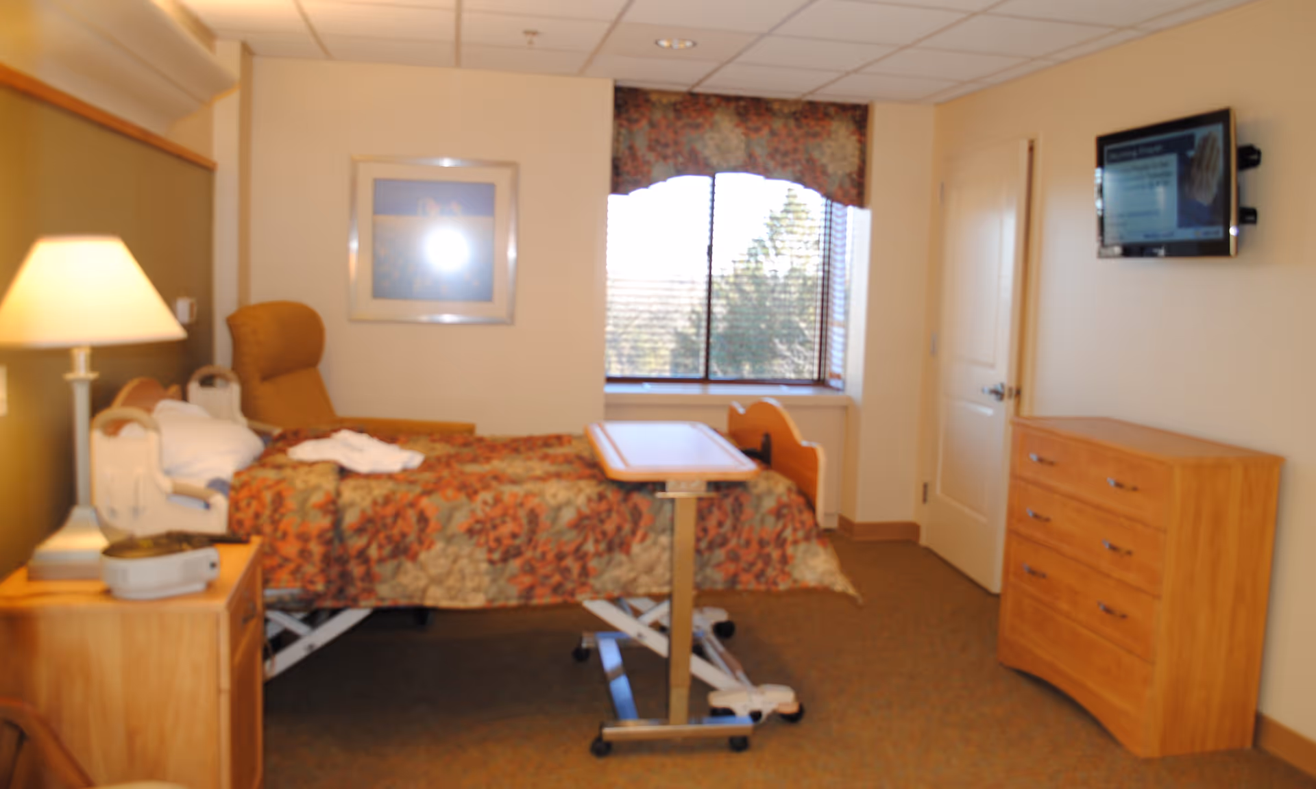 A senior living facility bedroom with a hospital-style adjustable bed covered with a floral bedspread. There is a wooden bedside table with a lamp and a device on it, a comfortable armchair next to the bed, a window with blinds and a floral valance, a wooden dresser, and a wall-mounted television.