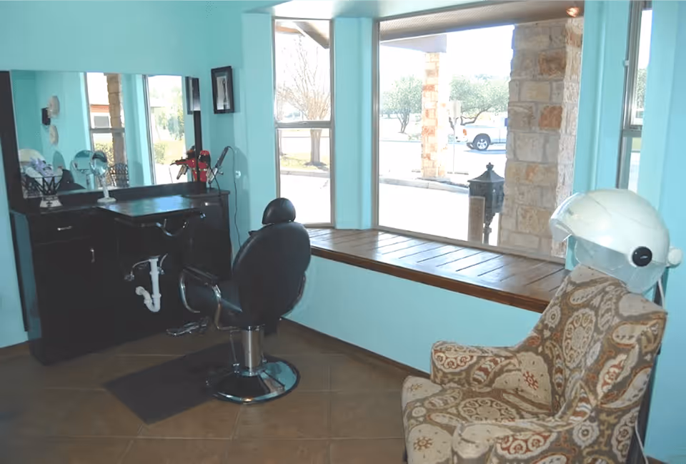 Interior view of a salon area in Mercy House Georgetown with a black salon chair in front of a mirror and counter, a patterned armchair with a white hair dryer hood, and large windows showing an outdoor view.