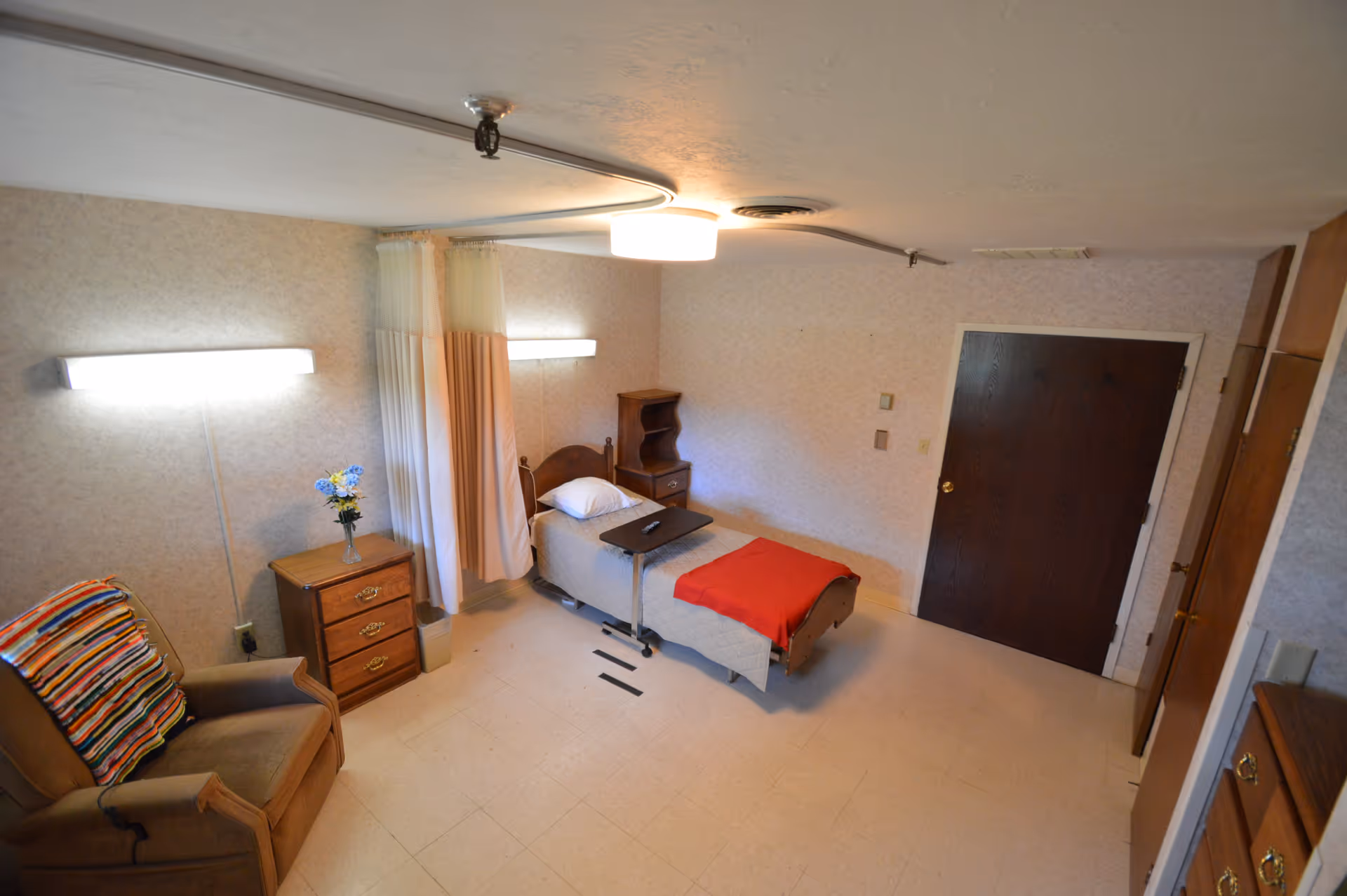 A small, simple room with a single bed covered with a beige blanket and a red throw. There is a wooden nightstand with a vase of flowers next to the bed, a brown armchair with a colorful striped blanket draped over it, and a small wooden cabinet. The room has beige walls, a ceiling light, and a dark wooden door. A privacy curtain is partially drawn around the bed.