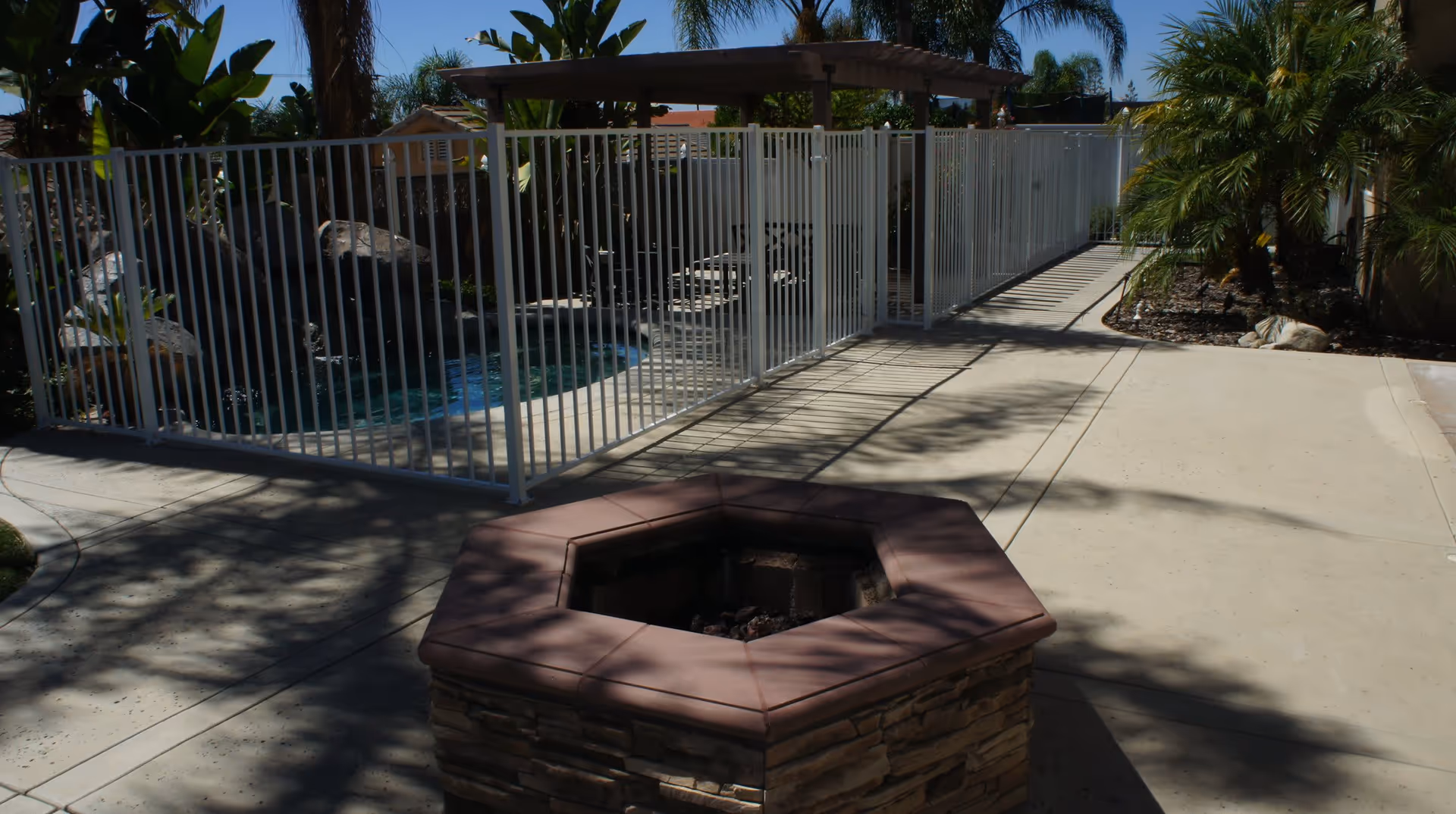 Outdoor patio area with a stone fire pit in the foreground, a concrete walkway, and a fenced swimming pool surrounded by tropical plants and palm trees under a clear blue sky.
