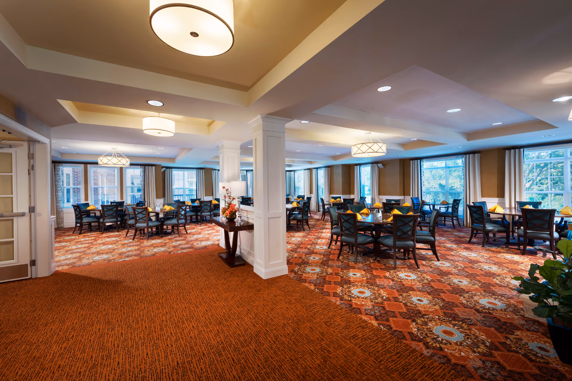 Spacious dining room with multiple round tables set with napkins, upholstered chairs, patterned carpet, large windows, and overhead lighting.