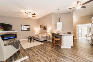 A spacious and modern living area featuring wood flooring, a ceiling fan, a wall-mounted electric fireplace, a beige armchair with a throw blanket, a sofa with cushions, a small side table with a lamp, and a kitchen island with two wooden stools. The kitchen area includes white cabinetry and stainless steel appliances, and there is a hallway leading to another room with large windows.