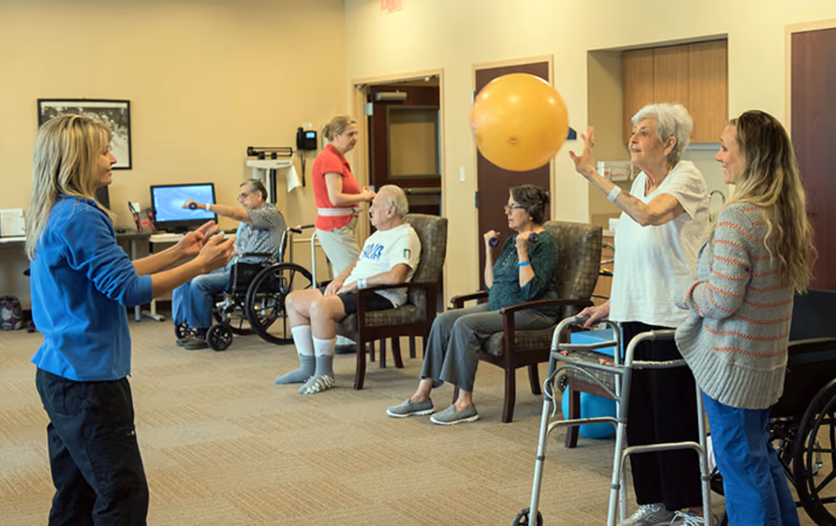 A group of elderly people and caregivers participate in a seated exercise activity in a spacious room. One elderly woman with a walker is tossing a large yellow ball to a caregiver, while others sit in chairs or wheelchairs, engaging in light exercise with small weights. The room has beige walls, carpeted floors, and wooden doors.