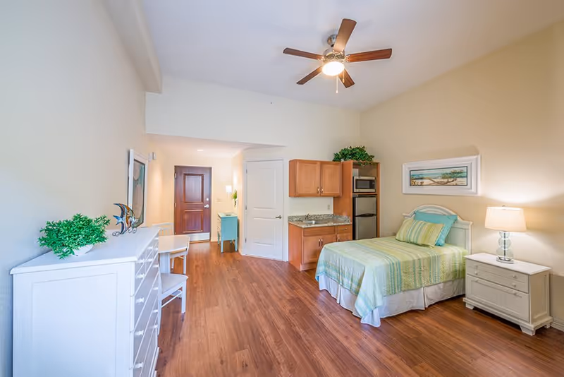 A bright and clean assisted living facility room with a single bed covered in green and blue striped bedding, a white nightstand with a lamp, a small kitchenette with a microwave and mini fridge, a white dresser with a plant and decorative fish sculpture, a ceiling fan with light, wooden flooring, and a wooden door at the far end.