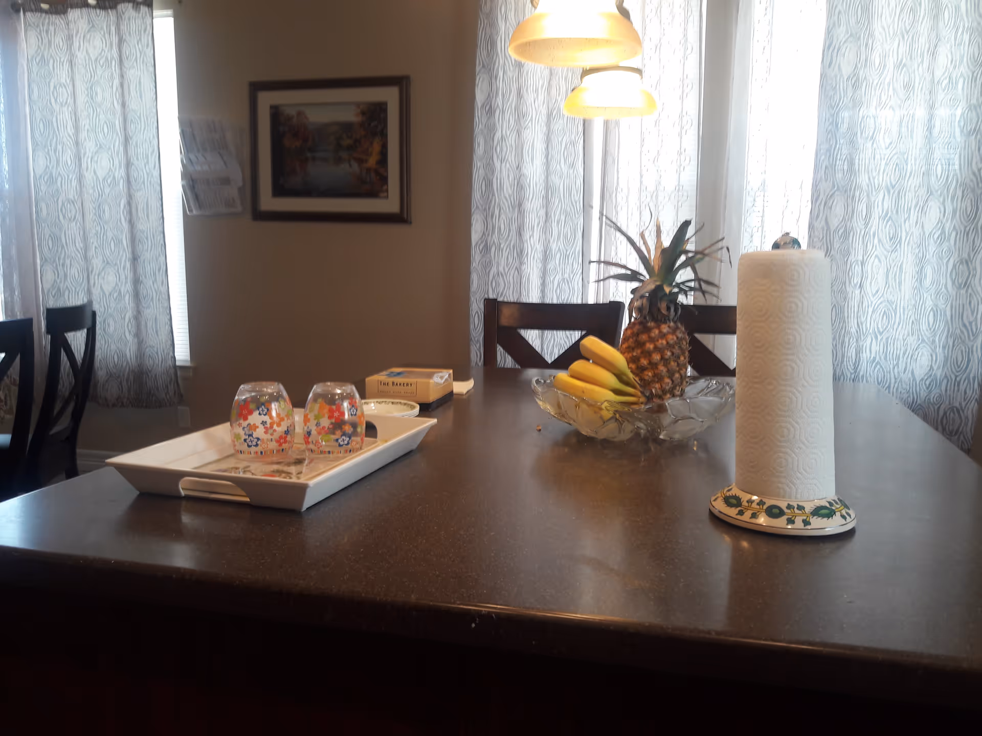 A dining area with a dark countertop table featuring a tray with two floral-patterned glasses, a bowl with bananas and a pineapple, a roll of paper towels on a decorative holder, and a box of tissues. The background shows chairs, patterned curtains, a framed picture on the wall, and hanging pendant lights.