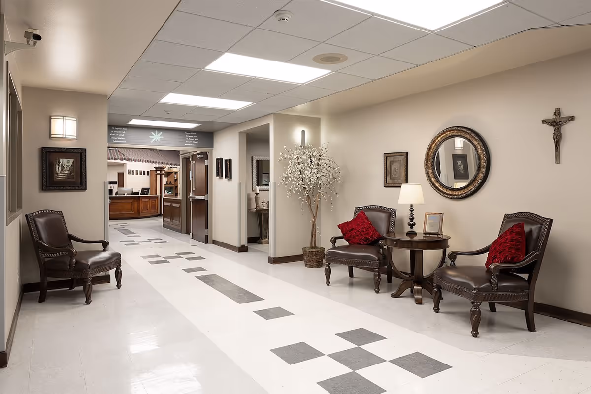 A clean and well-lit hallway in a senior living facility with polished tile floors featuring a gray and white pattern. On the right side, there are two dark wooden chairs with red cushions, a round wooden table with a lamp and a framed picture, a round decorative mirror, and a crucifix on the wall. A potted flowering tree is placed in a corner. On the left side, there is another dark wooden chair and a framed picture on the wall. At the end of the hallway, there is a reception desk with computer monitors and a sign above indicating directions to various rooms.