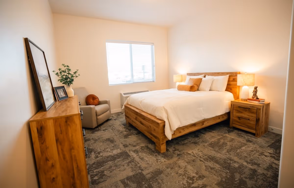 A cozy bedroom with a wooden bed frame and white bedding, two bedside tables with lamps, a wooden dresser with a framed picture and a vase with greenery, a comfortable armchair with a round cushion, and a window with blinds letting in natural light.