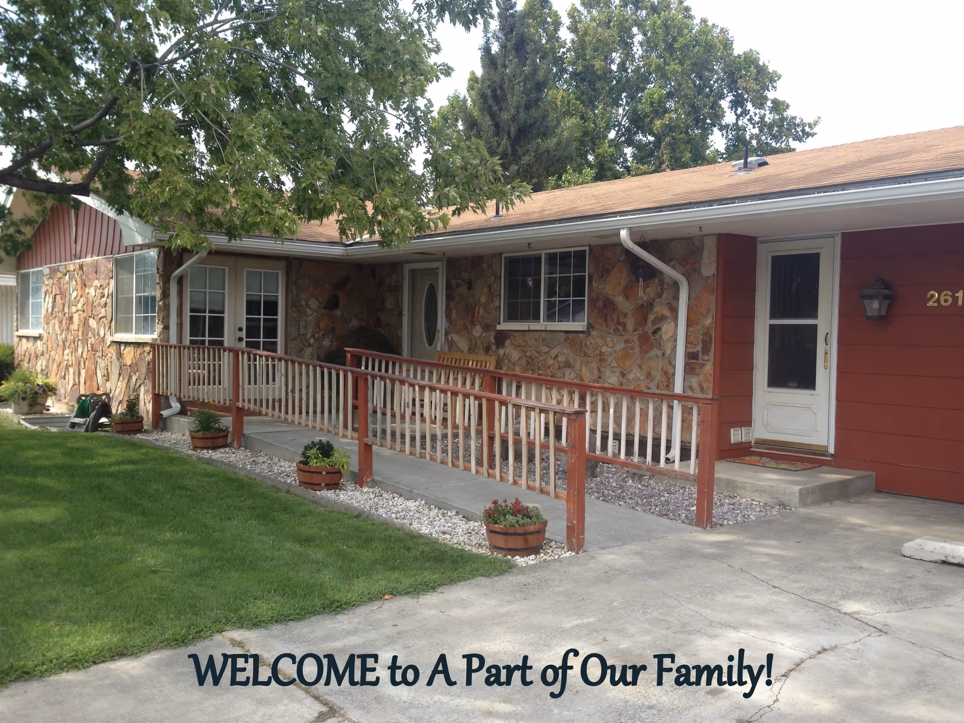 Exterior view of a single-story building with stone and red siding, featuring a wheelchair-accessible ramp with wooden railings, a concrete driveway, green lawn, and trees. The building has multiple windows and doors, and a welcome message saying 'WELCOME to A Part of Our Family!' is overlaid at the bottom of the image.