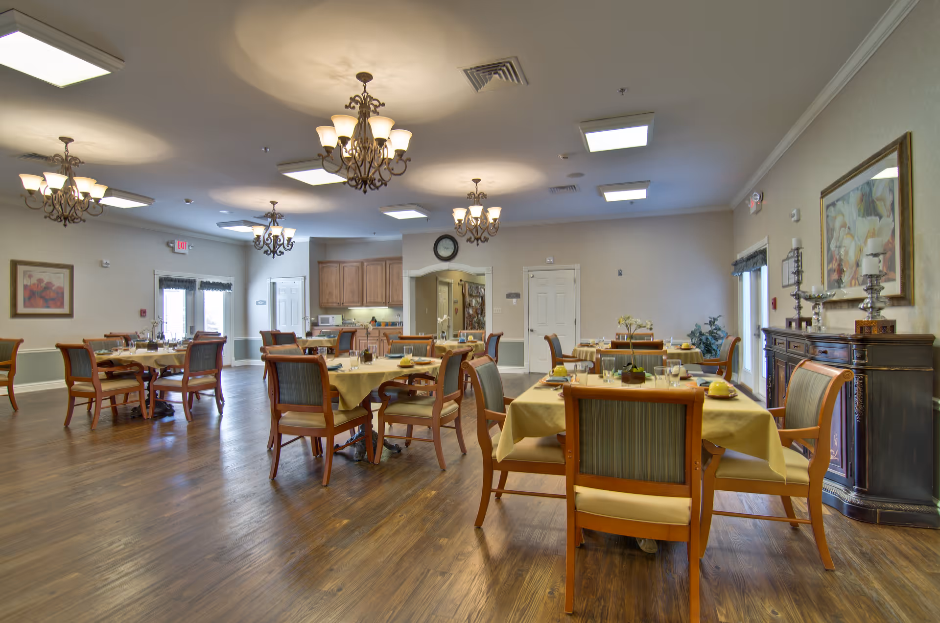 A spacious dining room in a senior living facility with multiple wooden tables covered with yellow tablecloths and set with plates, glasses, and utensils. The room features wooden chairs with green upholstery, hardwood floors, chandeliers hanging from the ceiling, and a sideboard with decorative items and a painting on the wall.