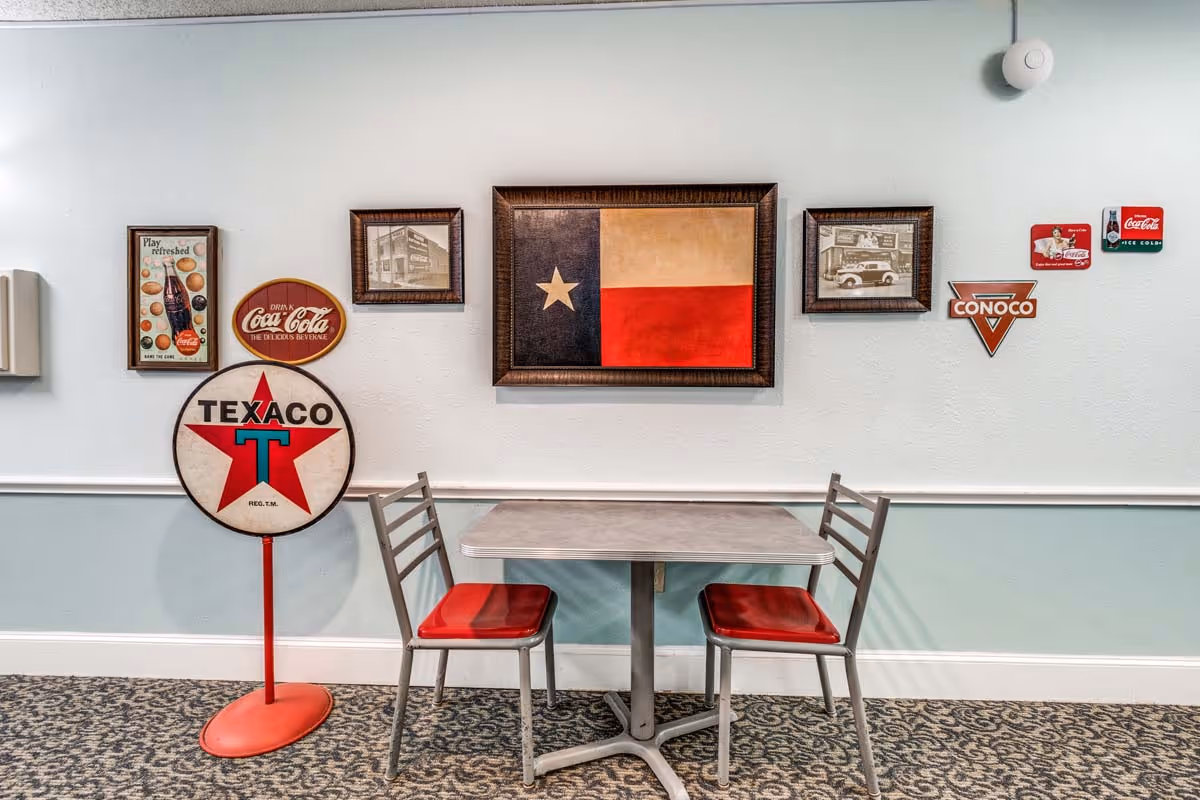 A small dining area with a square table and two metal chairs with red seats. The wall behind features vintage-style decorations including a framed Texas flag, old black and white photos, and retro Coca-Cola and Texaco signs. The floor is carpeted with a patterned design.