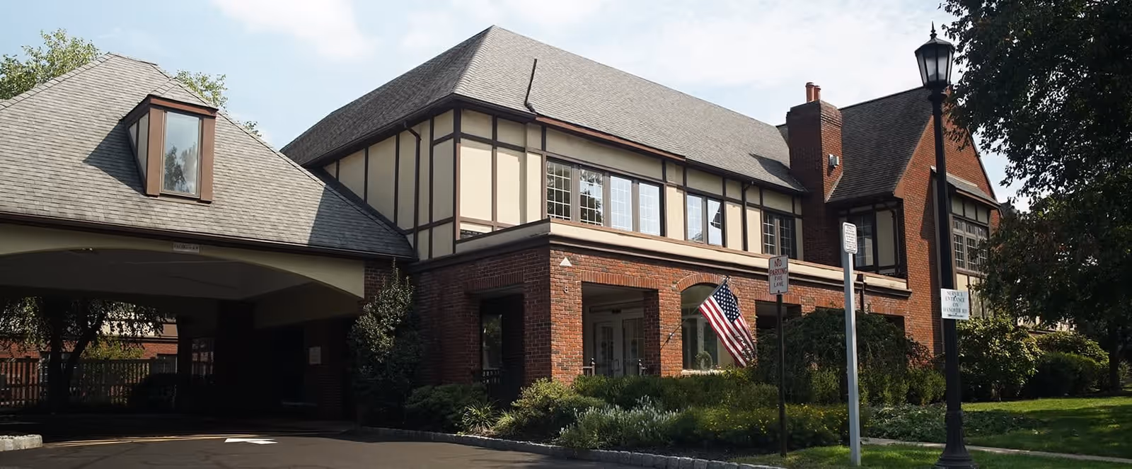 Exterior view of a two-story brick and timber senior living facility building with a covered driveway entrance, an American flag displayed near the entrance, and a street lamp with signage in front of the building surrounded by greenery.