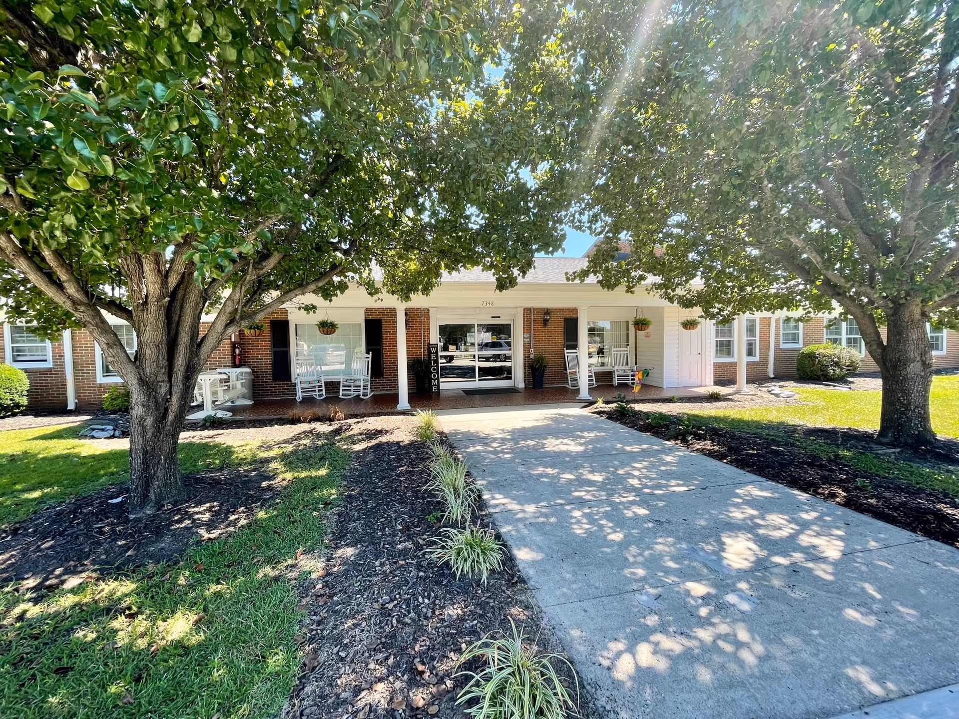 Front exterior view of Golden Years Nursing Home showing a single-story brick building with a covered porch. The porch has white rocking chairs and hanging plants. Two large trees flank the concrete walkway leading to the entrance, casting shadows on the ground.