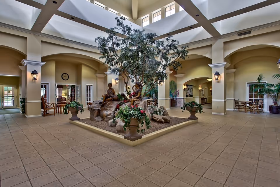 Spacious indoor common area with a large central planter featuring a tree, rocks, and two bronze statues of children. The room has high ceilings with skylights, beige tiled floors, and cream-colored walls with arches and columns. There are several seating areas with chairs and tables around the perimeter, and wall-mounted lantern-style lights provide illumination.