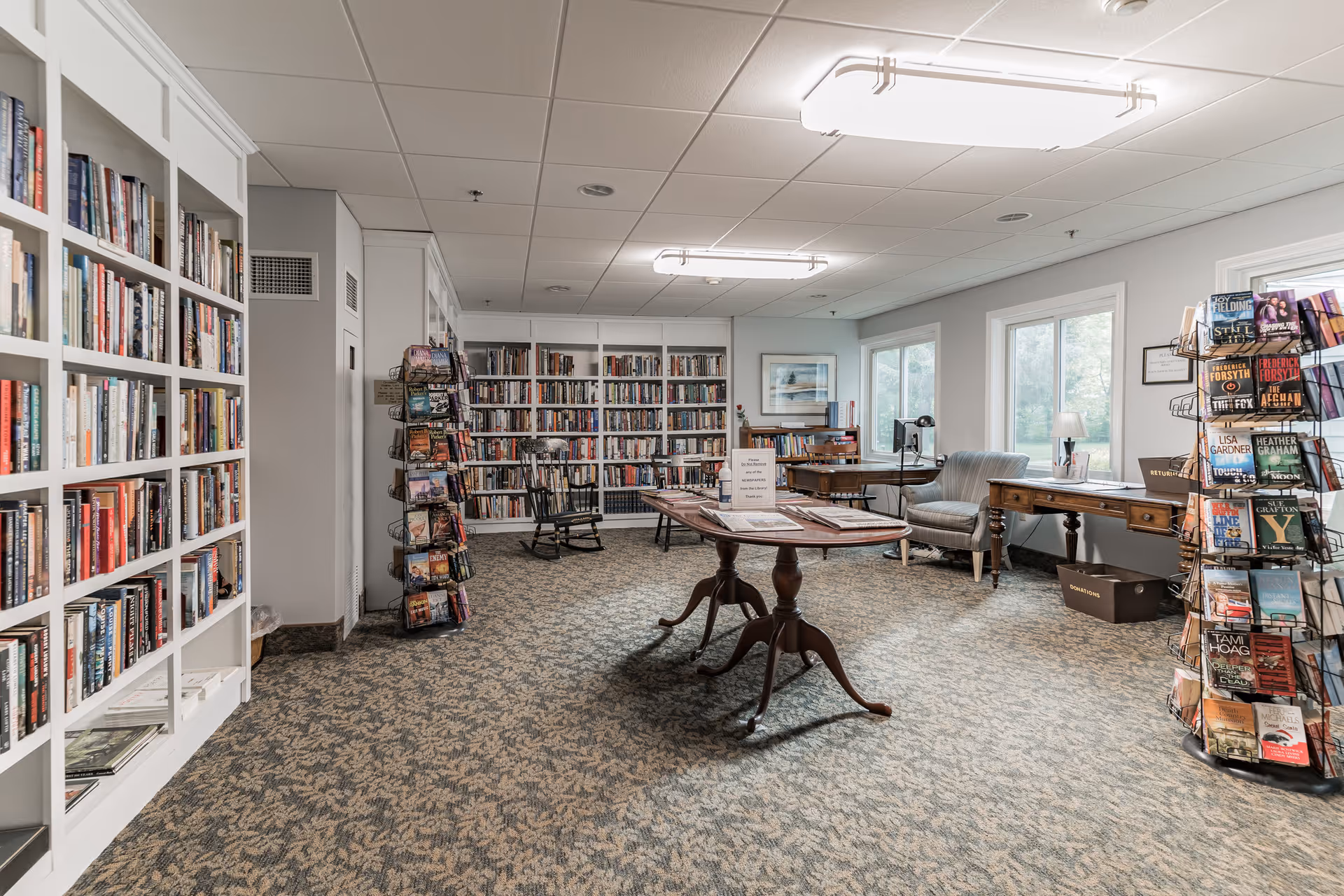 A well-lit library room with white bookshelves filled with books along the walls, two rotating racks with books or magazines, a wooden table in the center with reading materials, a rocking chair, a comfortable armchair, and large windows letting in natural light.