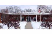 Front exterior view of Scenery Hill Healthcare and Rehabilitation Center building in winter with snow on the ground, leafless bushes lining the walkway, and an American flag on a flagpole near the entrance.