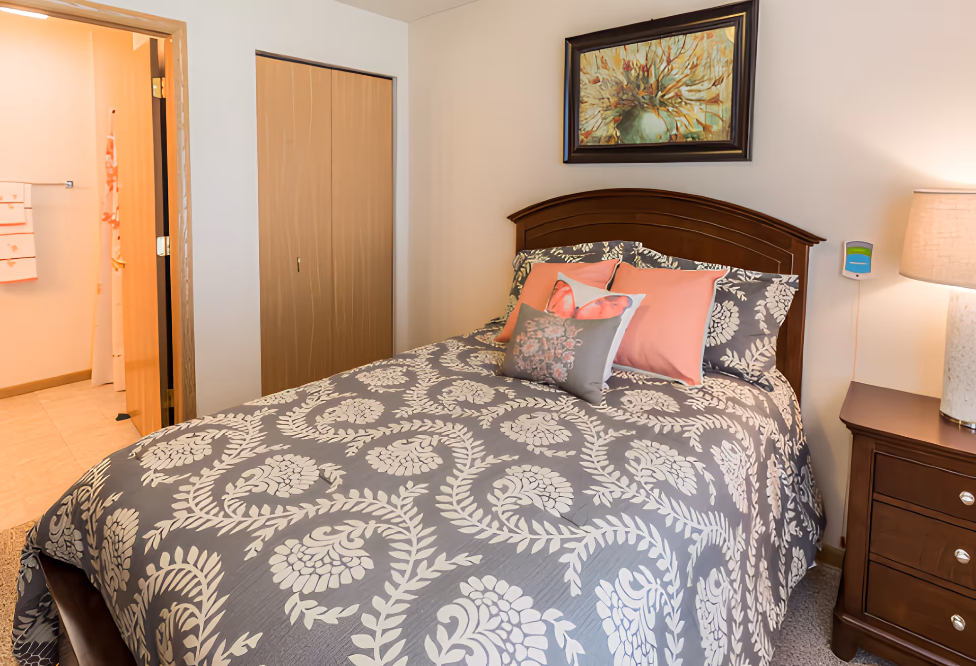 A cozy bedroom with a bed featuring a gray and white floral patterned bedspread and several decorative pillows in pink and gray. There is a wooden headboard and a nightstand with a lamp on the right side. A framed painting hangs on the wall above the bed. To the left, there is a doorway leading to a bathroom with visible towels and a shower curtain.