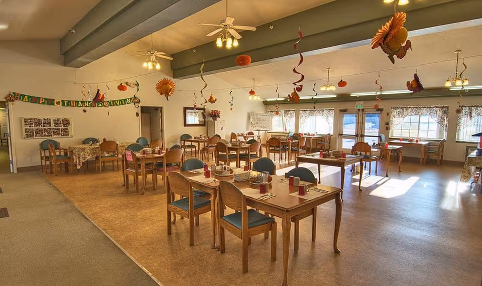 A spacious dining room decorated for Thanksgiving with hanging paper pumpkins, sunflowers, and spiral streamers. Several wooden tables with chairs are set with red cups, napkins, and utensils. Large windows let in natural light, and a 'Happy Thanksgiving' banner is displayed on the wall.