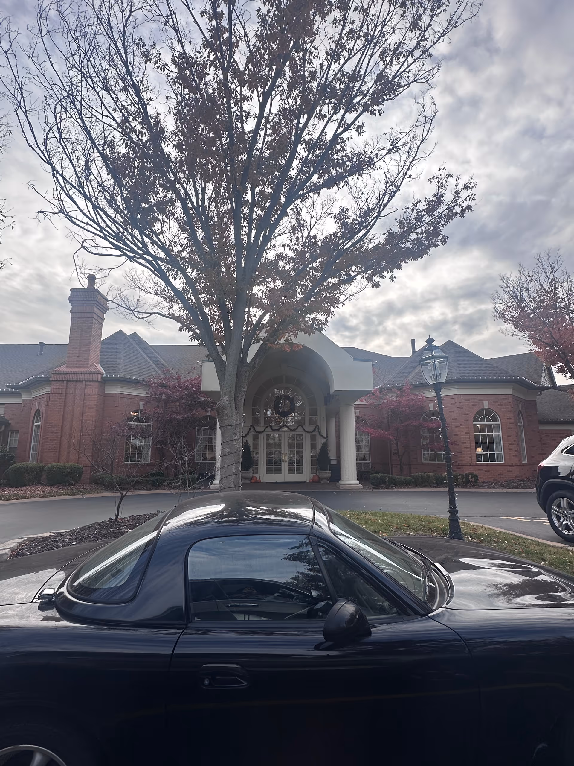Front exterior view of McKnight Place Extended Care building with a large tree in front, a black car parked nearby, and a cloudy sky overhead.
