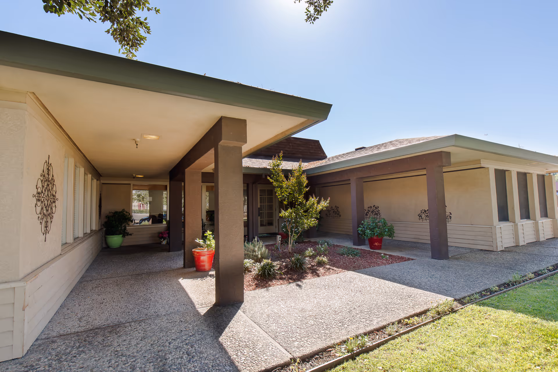 Exterior view of Windsor Elmhaven Care Center showing a covered walkway with brown pillars, beige walls, decorative wall art, potted plants, and a small landscaped area with shrubs and a tree under a clear blue sky.