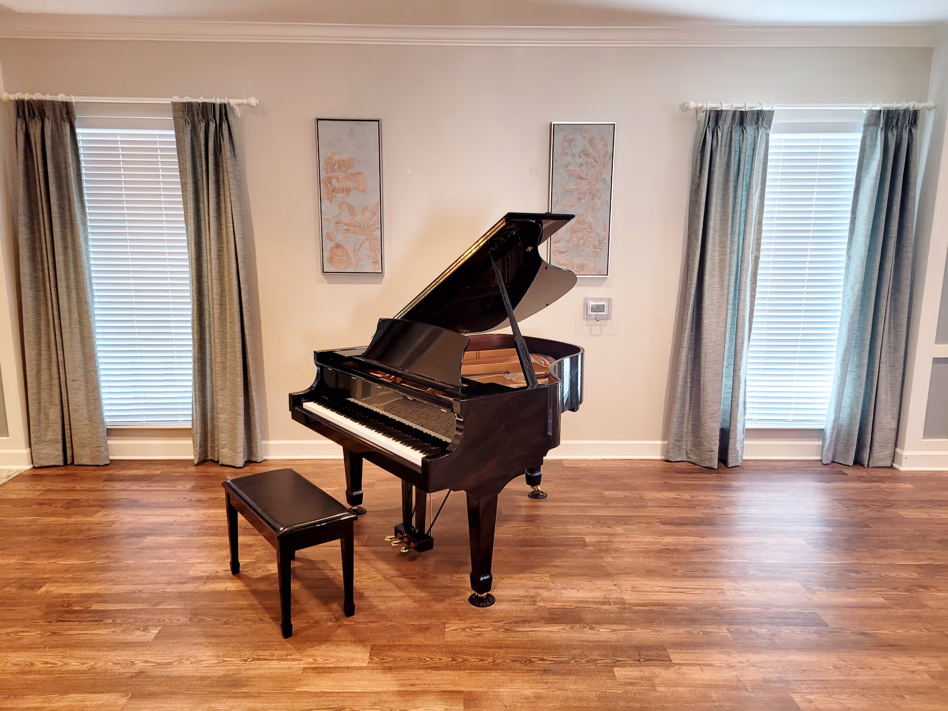 A black grand piano with an open lid and a matching bench is placed on a wooden floor in a room with two large windows covered by gray curtains and white blinds. Two framed artworks hang on the wall behind the piano.