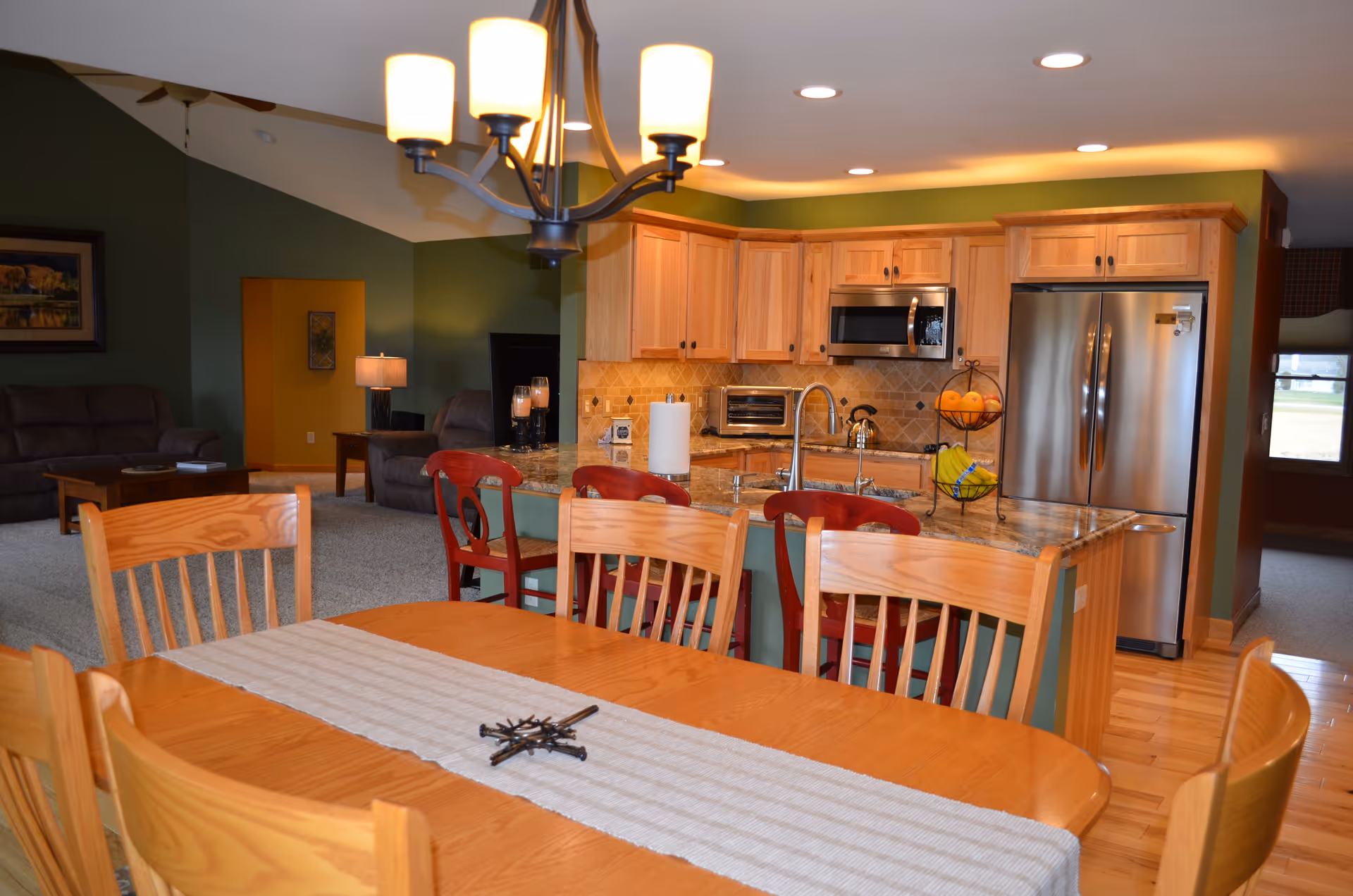 Open-plan dining area with a wooden table and chairs facing a modern kitchen island, stainless steel appliances, and a living room beyond.