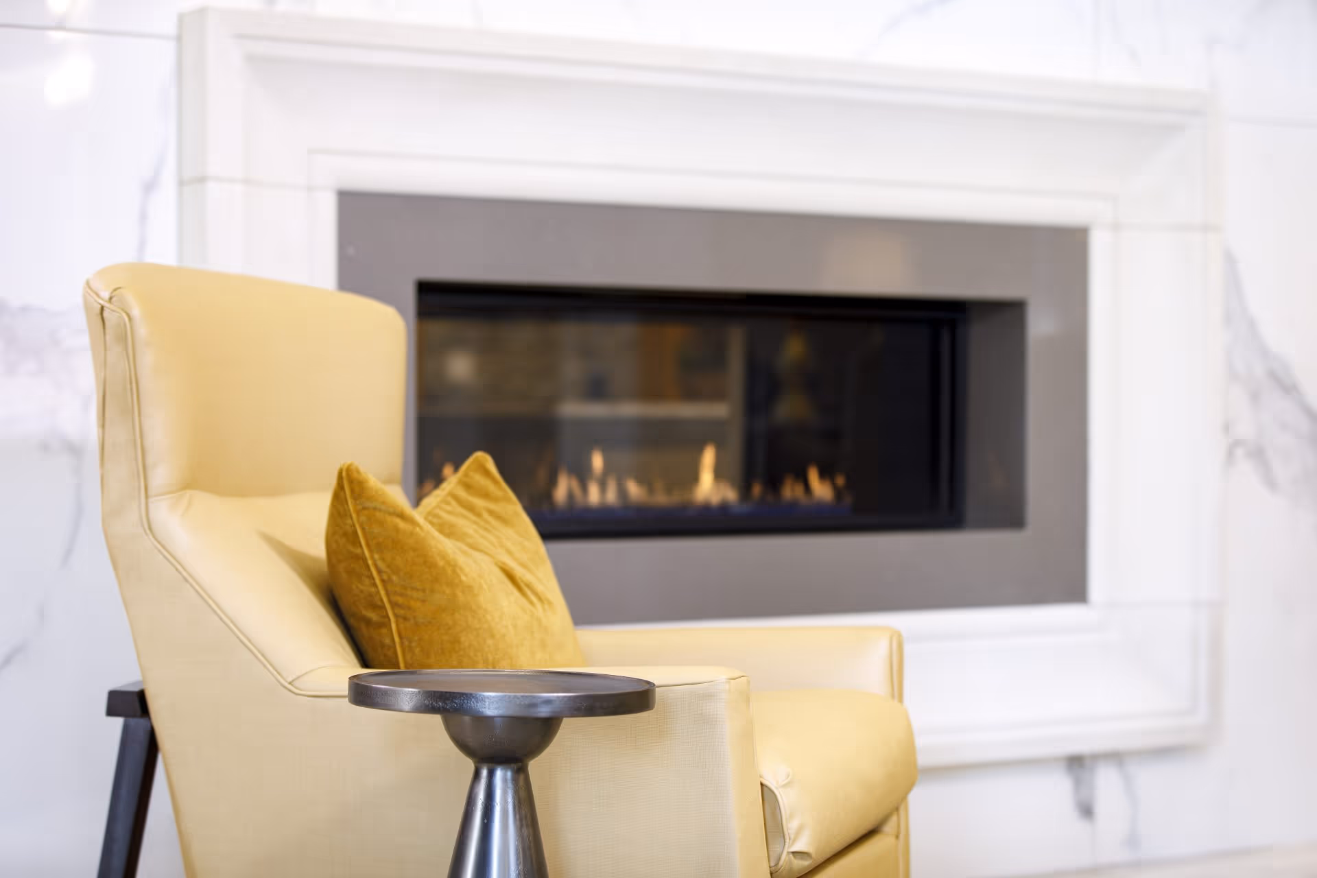 A cozy indoor scene featuring a light yellow armchair with a mustard yellow pillow, positioned next to a small round metallic side table. In the background, there is a modern rectangular gas fireplace set into a white marble wall.