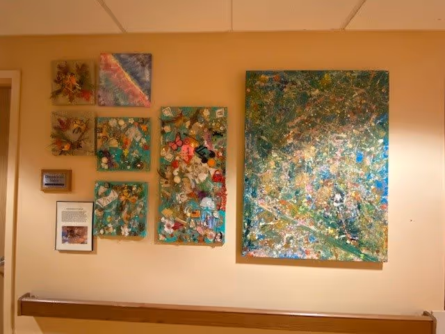 A hallway wall with a wooden handrail displaying several colorful abstract and mixed-media artworks.