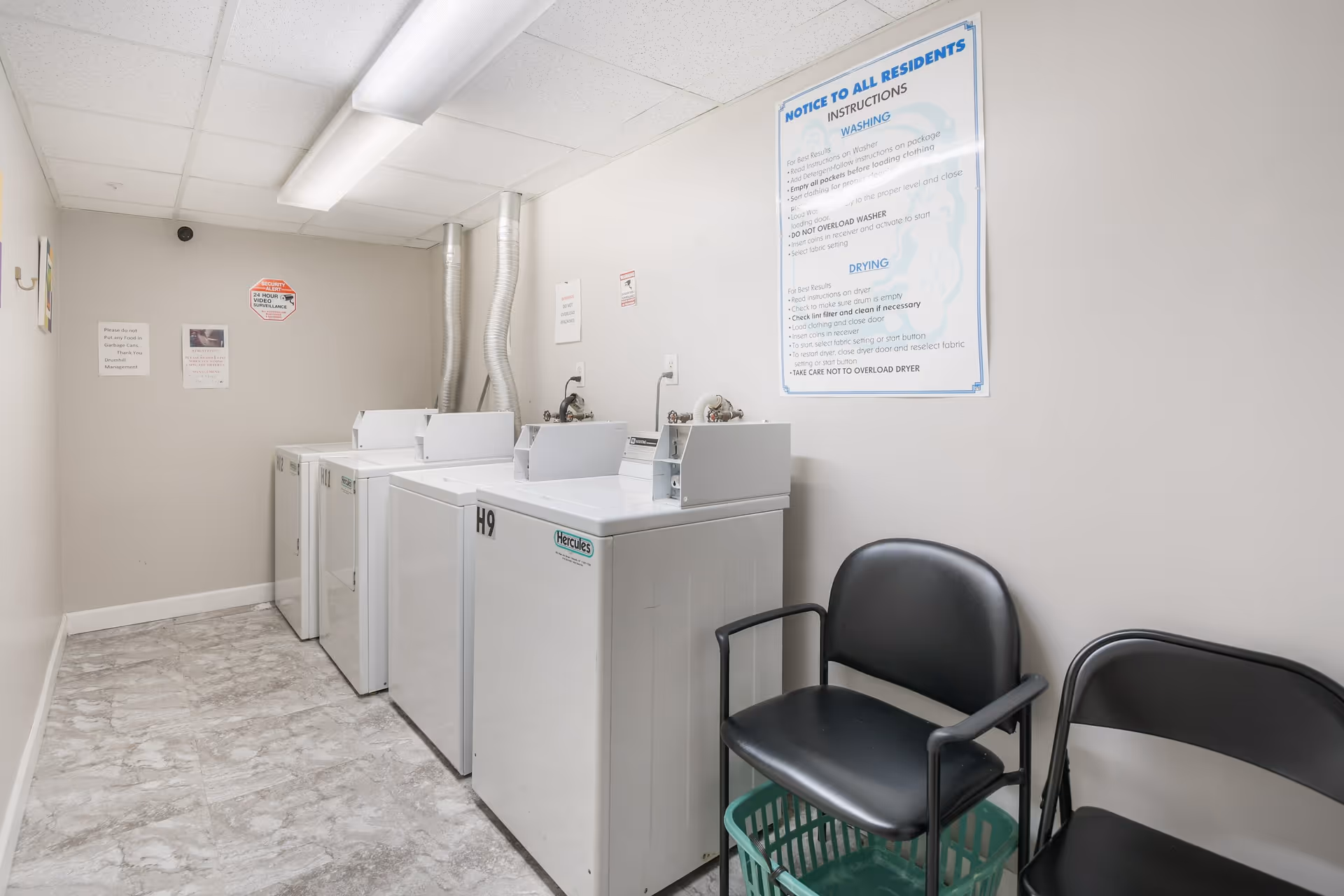 Laundry room with four white washing machines lined up against a beige wall. Above the machines is a large instructional sign for residents on washing and drying clothes. Two black chairs and a green laundry basket are placed next to the machines. The floor has a light gray tile pattern and the ceiling has white panels with fluorescent lighting.