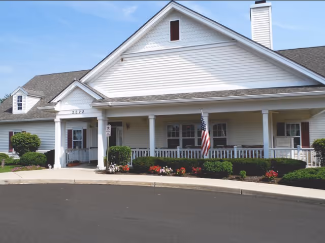Front entrance of a single-story senior living building with a covered porch, American flag, and landscaped flower beds.