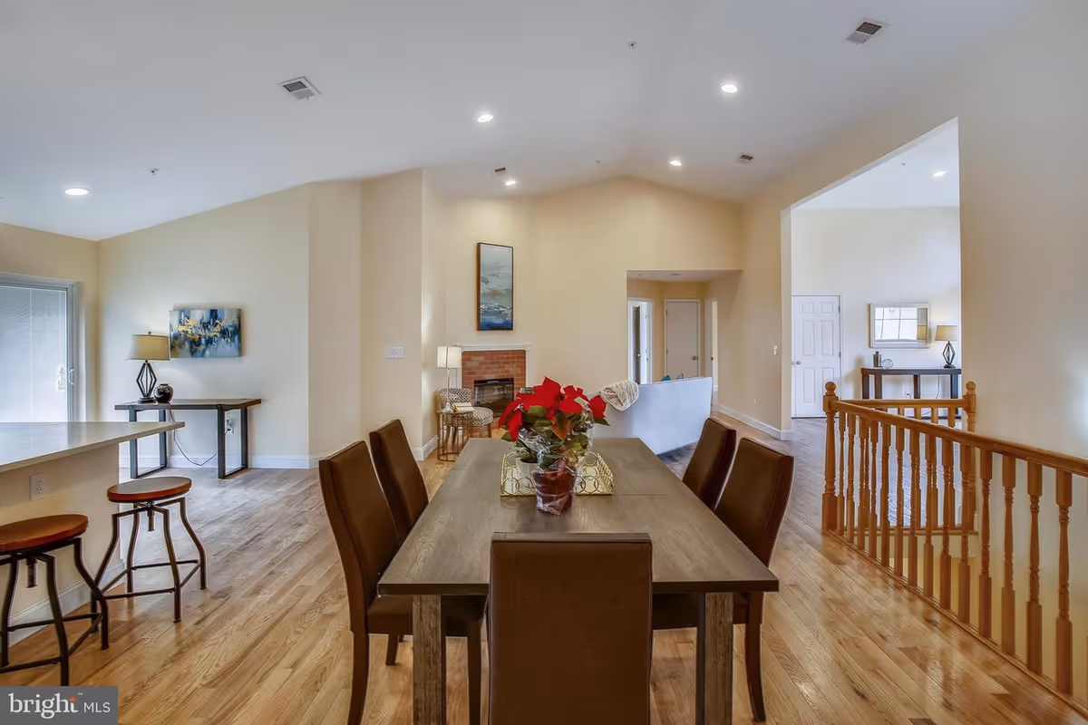 Interior view of a senior living facility showing a dining area with a wooden table and six brown chairs. A red flower centerpiece is on the table. To the left, there is a kitchen counter with two bar stools. In the background, there is a living room area with a sofa, a fireplace, and wall art. The floor is wooden, and the walls are painted light beige. There is a wooden railing on the right side and recessed lighting on the ceiling.