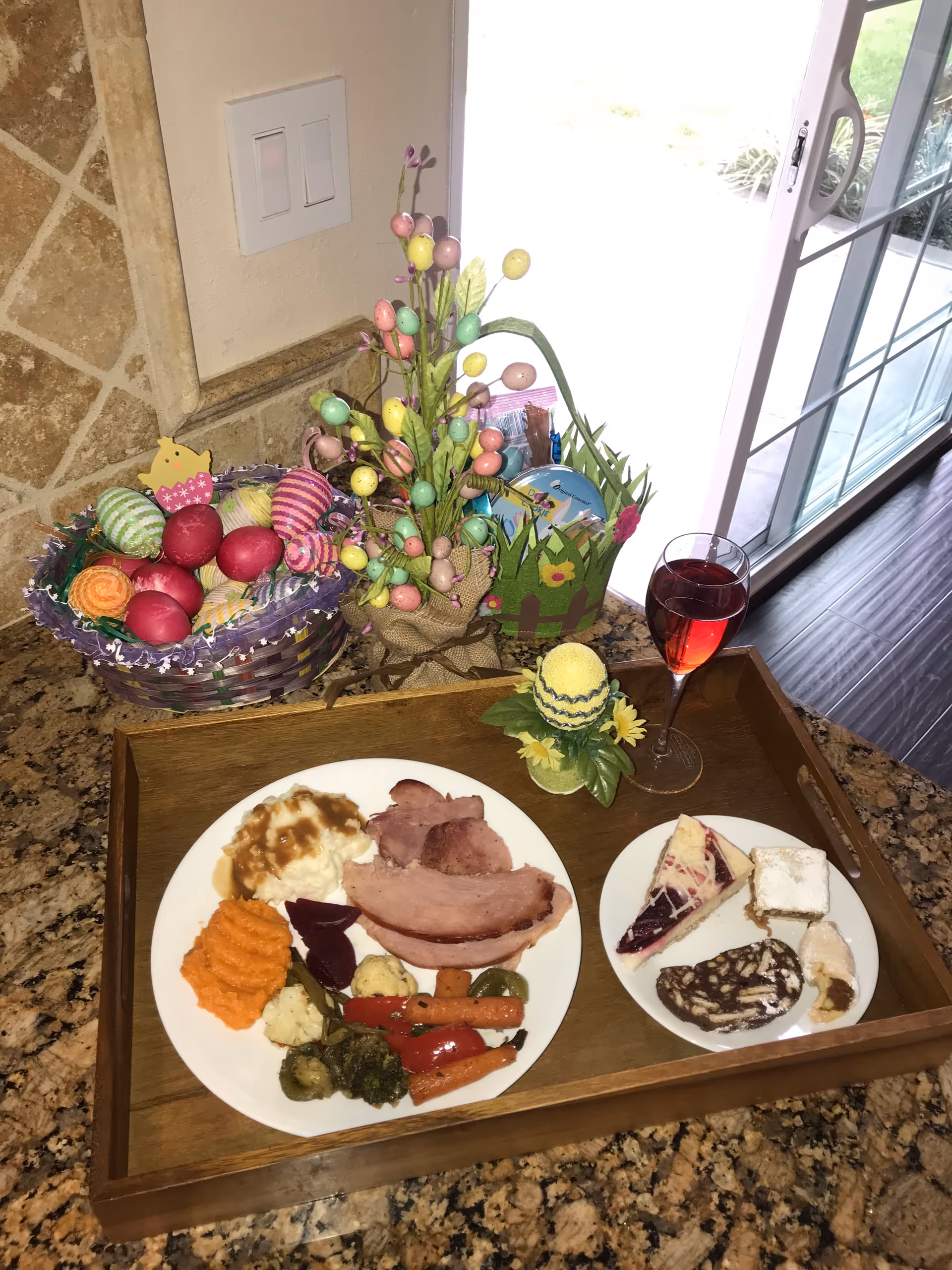 A wooden tray on a granite countertop holds a plate of food including mashed potatoes with gravy, ham slices, mashed sweet potatoes, and mixed vegetables. Next to it is a smaller plate with assorted desserts and a glass of red beverage. Behind the tray are Easter-themed decorations including a basket of colorful eggs, a small decorative tree with pastel eggs, and a green Easter basket. A sliding glass door is visible in the background.